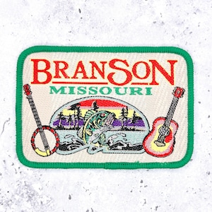 Puede incluir: Un parche bordado rectangular con borde verde sobre fondo crema. Presenta el texto rojo BRANSON y el texto verde MISSOURI. El diseño incluye un banjo a la izquierda, una guitarra a la derecha y una escena central de un pez saltando.