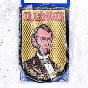 Op de afbeelding: Een rechthoekige geborduurde patch met een portret van Abraham Lincoln tegen een gouden achtergrond met ruitpatroon. De tekst ILLINOIS is in het rood bovenaan geborduurd en LAND OF LINCOLN in het goud onderaan.