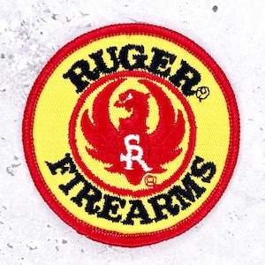 May include: A circular embroidered patch with a bright yellow background and a red border. The text RUGER FIREARMS is stitched in black around the centre. A red phoenix emblem is in the middle with a white SR logo superimposed on the bird.