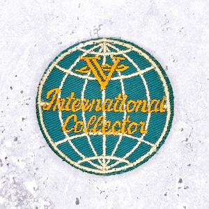 May include: A round, embroidered patch with a teal background and a white globe design. The text "International Collector" is in gold, along with a gold "V" emblem. The patch has a white border.