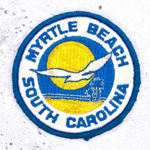 Pode incluir: Um emblema bordado circular com uma borda branca e o texto azul MYRTLE BEACH SOUTH CAROLINA. O centro retrata um sol amarelo, uma gaivota branca em voo, um horizonte oceânico azul e a silhueta de uma palmeira sobre um fundo azul.
