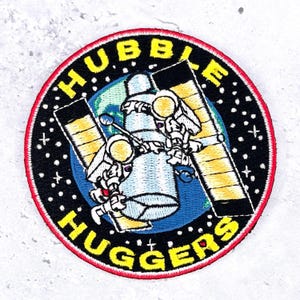 May include: A circular embroidered patch featuring a black space background with white stars. It depicts two astronauts in white suits hugging a central cylindrical structure, with yellow solar panels on either side. The text HUBBLE HUGGERS is in yellow.