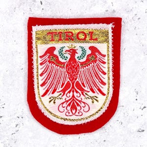 May include: A red and white embroidered patch with the word "TIROL" in gold lettering. The patch features a red eagle emblem with a green wreath, set against a white background, and a gold border.