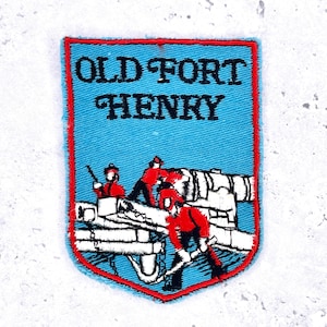 Może przedstawiać: Haftowana naszywka z napisem "OLD FORT HENRY" w kolorze czarnym na jasnoniebieskim tle. Naszywka ma czerwoną obwódkę i przedstawia żołnierzy i działo w kolorze białym i czerwonym.