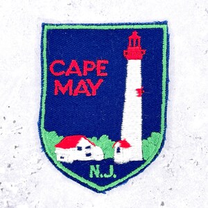 May include: A shield-shaped embroidered patch with a dark blue background. It features a white lighthouse with a red top, a small white building with a red roof, and green foliage. The text reads CAPE MAY in red and N.J. in green, with a green border.