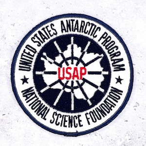 May include: A circular patch with a navy blue border and white text. The text reads "UNITED STATES ANTARCTIC PROGRAM" and "NATIONAL SCIENCE FOUNDATION". The center features a map of Antarctica and the letters "USAP" in red.