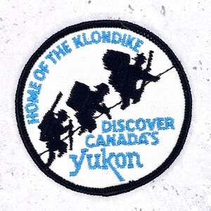 Könnte beinhalten: Ein kreisförmiger, gestickter Aufnäher mit weißem Hintergrund und schwarzem Rand. Der Aufnäher zeigt den Text "HOME OF THE KLONDIKE" und "DISCOVER CANADA'S YUKON" in Blau. Silhouetten von Goldsuchern sind ebenfalls enthalten.