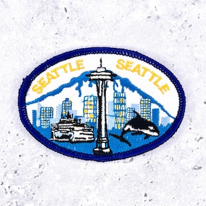 Op de afbeelding: Ovale patch met een Seattle stadsgezicht ontwerp. De patch bevat de Space Needle, gebouwen, bergen en een walvis. Het woord "SEATTLE" is in gele letters over de bovenkant gebogen, met een blauwe rand.