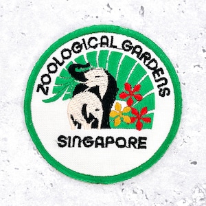 Może przedstawiać: Okrągła naszywka z haftem z zieloną obwódką. Zawiera napisy "ZOOLOGICAL GARDENS SINGAPORE" oraz ilustrację dwóch słoni, zielonych liści i czerwonych i żółtych kwiatów.