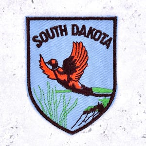 May include: A shield-shaped embroidered patch with a light blue background and a dark brown border. The text SOUTH DAKOTA is stitched in dark brown at the top. An orange and brown pheasant is depicted in flight above green grass and foliage.