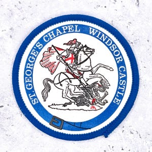 Könnte beinhalten: Ein runder Aufnäher mit blauem Rand und dem Text "ST. GEORGE'S CHAPEL WINDSOR CASTLE". In der Mitte ist ein Ritter auf einem Pferd zu sehen, der einen Drachen tötet, mit einem roten Kreuz auf dem Schild und der Flagge des Ritters.