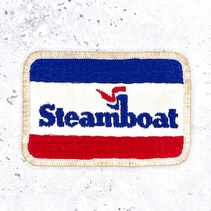 May include: A rectangular embroidered patch featuring a horizontal tricolor design of blue, white, and red. The word Steamboat is stitched in blue across the white centre stripe, with a small red and blue graphic element above the letter m.