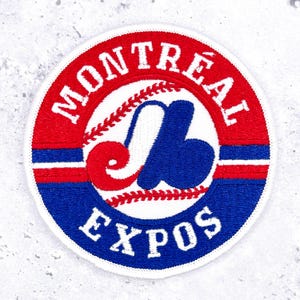 May include: A circular embroidered patch featuring the Montreal Expos baseball logo. The design includes a red top section with white text reading MONTREAL, a blue bottom section with white text reading EXPOS, and a central stylised blue and red M.