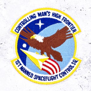 May include: Embroidered patch with a blue and yellow border, featuring a brown eagle with white head and a rocket. Text reads "CONTROLLING MAN'S HIGH FRONTIER" and "1ST MANNED SPACEFLIGHT CONTROL SQ."