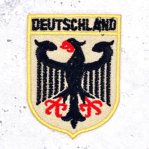 Peut inclure: Un écusson brodé en forme de bouclier portant le mot DEUTSCHLAND en lettres capitales noires en haut. Sous le texte se trouve un aigle noir au bec et aux griffes rouges centré sur un fond beige, le tout bordé d'un contour jaune cousu.