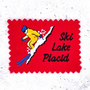May include: A red patch with a skier in a yellow suit and blue skis, skiing down a white slope. The text "Ski Lake Placid" is in black.