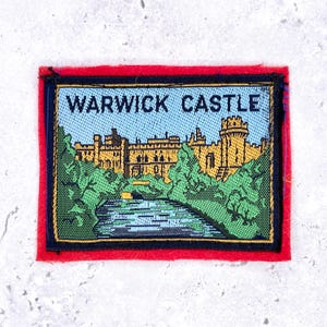 May include: A rectangular embroidered patch featuring a woven scene of Warwick Castle in yellow, set against a light blue sky and surrounded by green trees. A blue river flows in the foreground. The text WARWICK CASTLE is stitched in black.