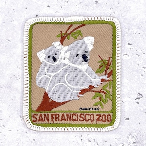 May include: Embroidered patch featuring two gray koalas perched on a brown tree branch with green leaves. The patch has a tan background and a green border with the text "SAN FRANCISCO ZOO". The patch is rectangular with a white stitched border.