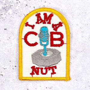 Puede incluir: Parche bordado con fondo blanco, borde amarillo y texto rojo que dice "I AM COB NUT". Un micrófono azul está centrado sobre una base gris y circular.