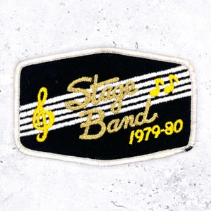 May include: Black embroidered patch with the words "Stage Band 1979-80" in gold lettering. The patch features a treble clef and musical notes in yellow, with white stripes across the center.