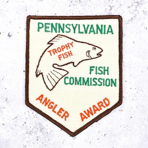 May include: A shield-shaped embroidered patch with a cream background and brown border. It features a brown outline of a fish, the text PENNSYLVANIA, TROPHY FISH, FISH COMMISSION, and ANGLER AWARD in green and orange thread on the fabric surface.