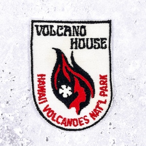 Pode incluir: Um patch branco com acabamento preto com o texto "VOLCANO HOUSE" e "VOLCANOES NAT'L PARK". Um design de chama vermelha e preta está no centro, com "HAWAII" em texto vermelho. O patch tem formato de escudo.