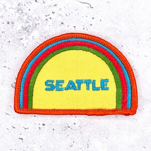 Puede incluir: Un parche bordado en forma de semicírculo con un centro amarillo brillante que presenta la palabra SEATTLE bordada en azul claro. El parche está bordeado por bandas concéntricas verdes, azul claro y rojas, con un borde grueso cosido en naranja.