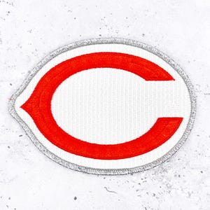 Peut inclure: Écusson brodé ovale blanc avec un "C" rouge et une bordure argentée. Le patch est fait d'un tissu texturé et d'un bord cousu, adapté aux vêtements ou accessoires.