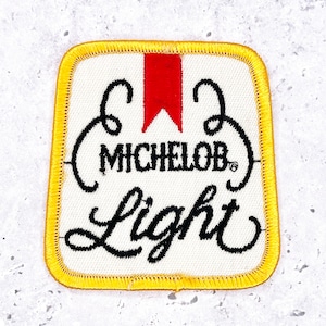 May include: Square patch with a white background, yellow border, and the words "Michelob Light" in black script. A red accent is at the top. The patch is likely for a jacket or bag.