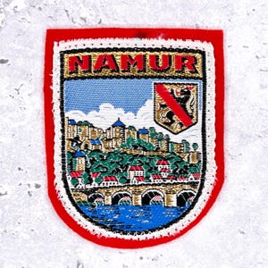 Pode incluir: Remendo retangular com borda vermelha e design bordado detalhado. O remendo apresenta a palavra "NAMUR" no topo, uma paisagem urbana e um brasão de armas. O design inclui um céu azul, um rio e uma ponte.