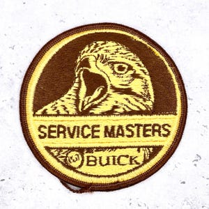 Puede incluir: Un parche redondo bordado con un borde marrón y un diseño amarillo y marrón. El parche presenta la cabeza de un águila sobre las palabras "SERVICE MASTERS" y "BUICK".