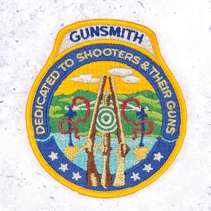 May include: A shield-shaped embroidered patch with a yellow border. The text reads GUNSMITH and DEDICATED TO SHOOTERS & THEIR GUNS. The design features two rifles, a green target, red ribbons, and a landscape with blue water and green hills.
