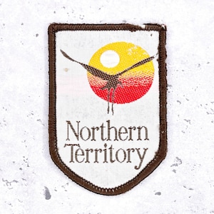 May include: A rectangular patch with a brown border and the words "Northern Territory" in brown text. The patch features a stylized image of a bird in flight against a sunset with yellow, red, and brown colors.