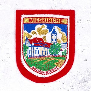 May include: A rectangular patch with a red border and a detailed embroidered image. The image features a church with a red roof, blue sky, and the text "WIESKIRCHE" and "WALLFAHRTSORT". The patch has a white background.