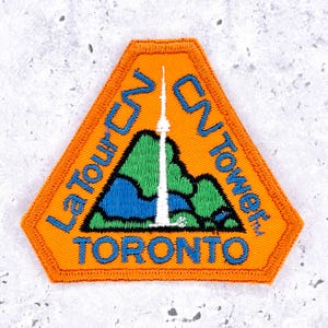 Op de afbeelding: Oranje, driehoekige patch met de CN Tower in Toronto. De patch heeft blauwe tekst met de tekst "La Tour CN Tower, Toronto". De CN Tower is afgebeeld in wit met groene en blauwe accenten.