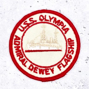 May include: Circular patch with a red border and text reading "U.S.S. OLYMPIA ADMIRAL DEWEY FLAGSHIP." The center features a detailed embroidered image of a ship in beige and white.