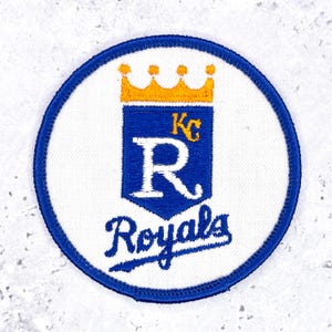 May include: A circular embroidered patch featuring the Kansas City Royals logo. The design includes a yellow crown above a blue shield with a white letter R and the letters KC in yellow. The word Royals is stitched in blue below the shield.