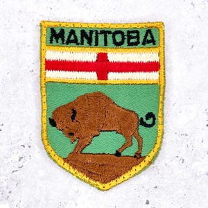 May include: Embroidered patch featuring the word "MANITOBA" above a flag design with a red cross and white stripes. Below, a brown bison stands on a green background. The patch has a yellow border.