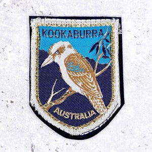 Könnte beinhalten: Schildförmiger Aufnäher mit einer Illustration eines Kookaburra-Vogels. Der Aufnäher hat einen blauen Hintergrund mit den Worten "KOOKABURRA" und "AUSTRALIA" in Weiß. Der Aufnäher ist mit Gold und Weiß verziert.