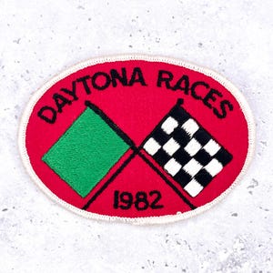 May include: Oval-shaped patch with a red background and white stitched border. The patch features the words "DAYTONA RACES" in black, arched across the top. Two flags are depicted: a green flag and a black and white checkered flag. The year "1982" is printed at the bottom.