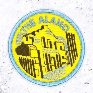 Könnte beinhalten: Ein runder gestickter Aufnäher mit leuchtend gelbem Hintergrund und hellblauem Rand. Das Design zeigt eine dunkelbraune Illustration des Alamo-Missionsgebäudes mit dem in Hellblau oben bogenförmig aufgestickten Text THE ALAMO.