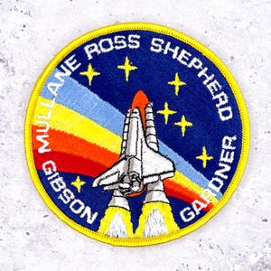 May include: Embroidered patch featuring a space shuttle design with the names MULLANE ROSS SHEPHERD GIBSON GARDNER. The patch has a blue background with yellow stars and a rainbow-colored arc. The shuttle is white and gray.