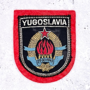 May include: A vintage Yugoslavia patch with a red border. The patch features the word "YUGOSLAVIA" above a central emblem with a red star, flames, and the date "29-XI-1943". The emblem is surrounded by gold and blue details.