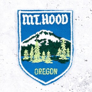 Könnte beinhalten: Gestickter Aufnäher mit dem Mt. Hood mit schneebedecktem Gipfel, grünen Bäumen und dem Text "MT. HOOD OREGON" in Weiß und Grün auf blauem, schildförmigem Hintergrund mit weißem Rand. Ideal für Jacken, Taschen oder Reisesouvenirs.