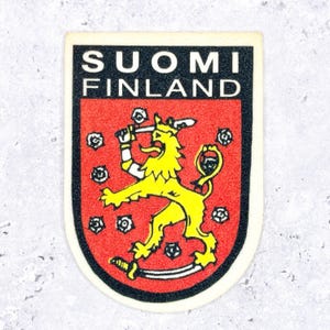 May include: A shield-shaped patch featuring the coat of arms of Finland with a yellow lion on a red background, surrounded by white roses. The text SUOMI FINLAND is printed in white on a black horizontal bar at the top of the patch design.