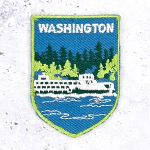 Può includere: Una toppa ricamata a forma di scudo con sfondo blu e la scritta WASHINGTON in bianco nella parte superiore. Il disegno raffigura un traghetto bianco su acqua blu, davanti a una foresta di conifere nei toni del verde scuro e chiaro.