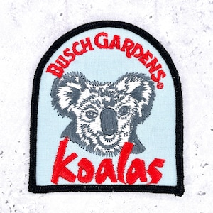 May include: An embroidered patch with a light blue background and a black border. It features a gray and white koala head in the center. The text BUSCH GARDENS is arched in red above the koala, and the word Koalas is printed in red below it.