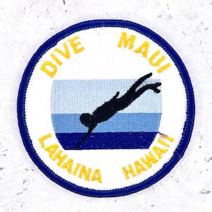 May include: A circular embroidered patch with a white background and a dark blue border. The centre features a black silhouette of a diver swimming through horizontal bands of light blue and dark blue. Yellow text reads DIVE MAUI LAHAINA HAWAII.