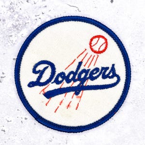 Op de afbeelding: Een ronde geborduurde patch met een witte achtergrond en een blauwe rand. Het woord Dodgers is in blauw schrift in het midden geborduurd, vergezeld van een rode honkbalafbeelding met bewegingslijnen die erachter zijn gestikt.