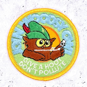 May include: A circular embroidered patch with a yellow border. It features a brown owl wearing a green hat with a red feather, holding a white tube. The background is light green with an orange sun and the text GIVE A HOOT DON'T POLLUTE.